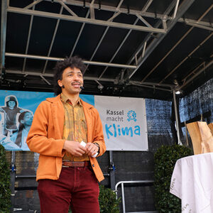 Der Moderator auf der Bühne, im Hintergrund das Banner das Klimaschutzpreises. Rechts auf einem Tisch stehen die Skulpturen