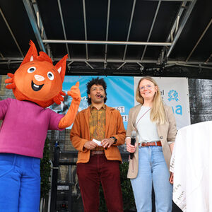 Ein Baumhörnchen-Kostüm mit blauer Hose und lila T-Shirt steht auf der Bühne und zeigt beide Daumen nach oben. Daneben der Moderator David Zabel und Lena Chmielewski.