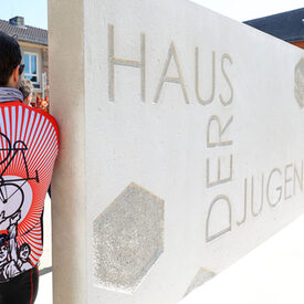 Zementblock mit Inschrift "Haus der Jugend". Daneben lehnt sich ein Mann mit orangefarbenen Shirt an.