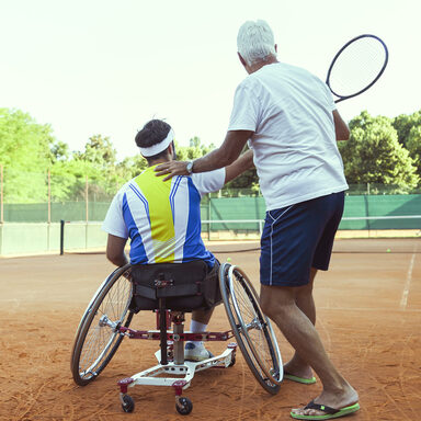 Tennislehrer bringt Mann im Rollstuhl das Tennisspielen bei.