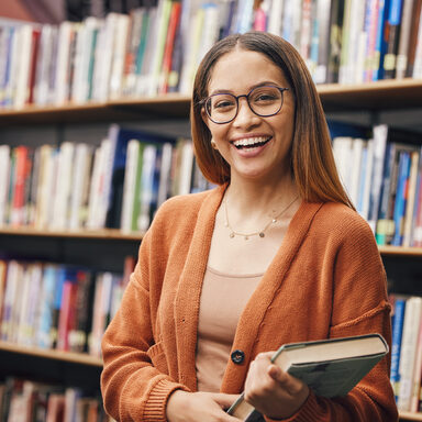 Eine junge Studentin steht vor einem Bücherregal und hat ein Buch in der Hand. Sie lächelt.