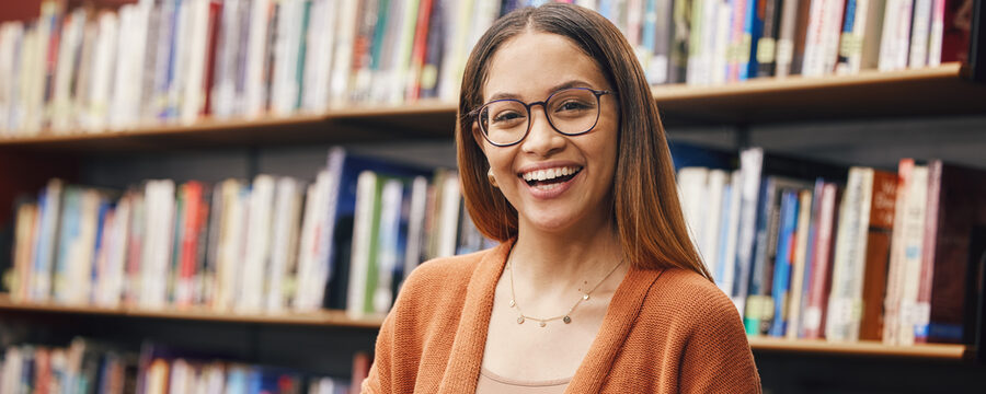 Eine junge Studentin steht vor einem Bücherregal und hat ein Buch in der Hand. Sie lächelt.