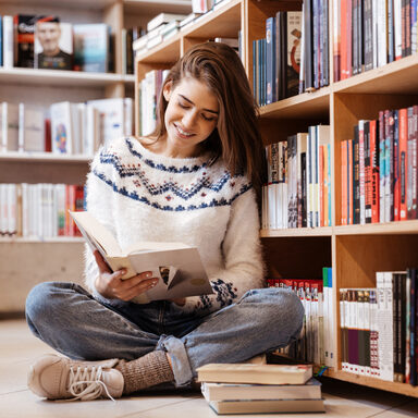 Eine junge Frau liest ein Buch, während sie in der Bücherei im Schneidersitz auf dem Boden sitzt.