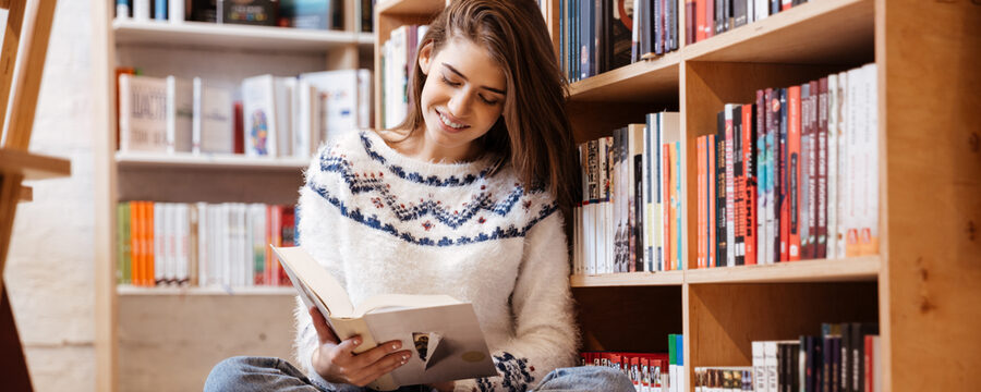 Eine junge Frau liest ein Buch, während sie in der Bücherei im Schneidersitz auf dem Boden sitzt.
