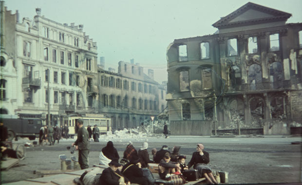 Menschen sitzen auf Koffern, im Hintergrund zerstörte Gebäude.