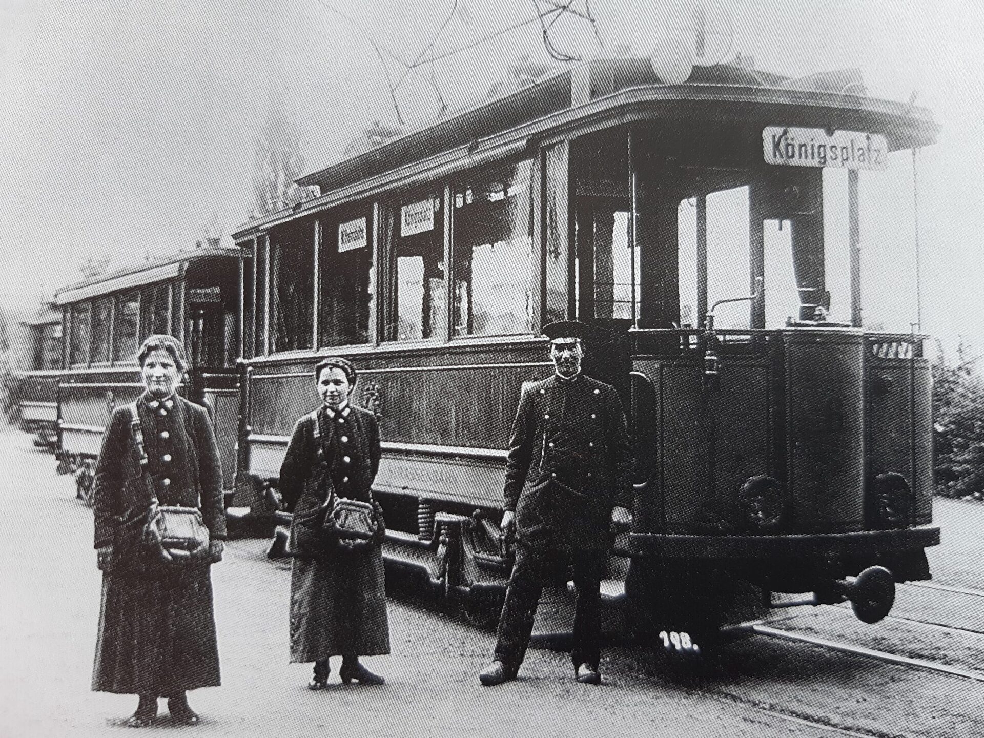 Fahrer und Frauen mit Umhängetaschen vor einer Stra0enbahn mit dem Schild Königsplatz