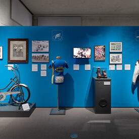 Eine Wand, an der der Radsport und Rollerderbys vorgestellt werden. Für beide ist jeweils ein Oberteil und ein Sportgerät ausgestellt.