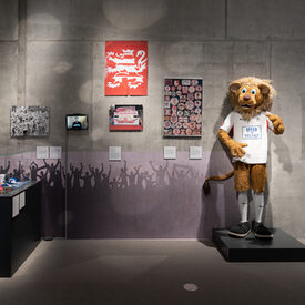 Eine Ecke mit einer Vitrine über die Fankultur beim Sport und an der Wand daneben hängen Bilder und ein Video über den KSV Hessen-Kassel. Ein Kostüm des Maskottchen „Totti“ (Löwe) steht daneben.