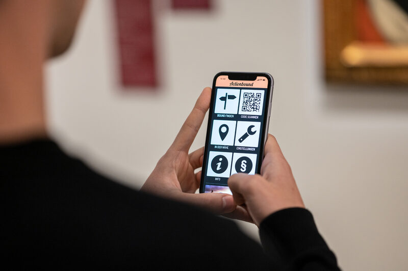 Ein Handy mit der App Actionbound in der Hand eines Besuchers des Museums
