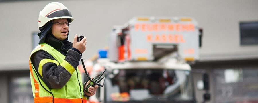 Ein Feuerwehrmann spricht in ein Funkgerät und steht vor einem Feuerwehrauto.